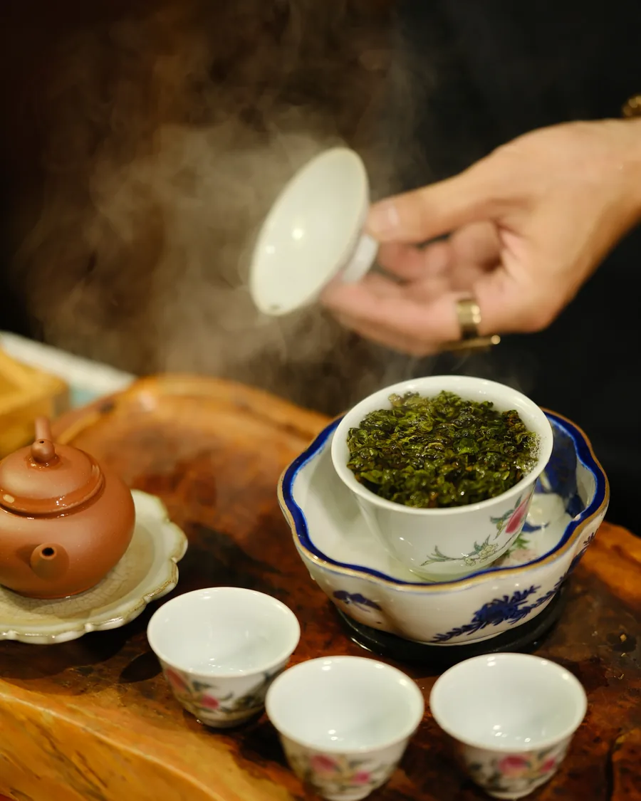 A hand lifting the lid from a gaiwan heaped with green leaves as steam rises over waiting cups
