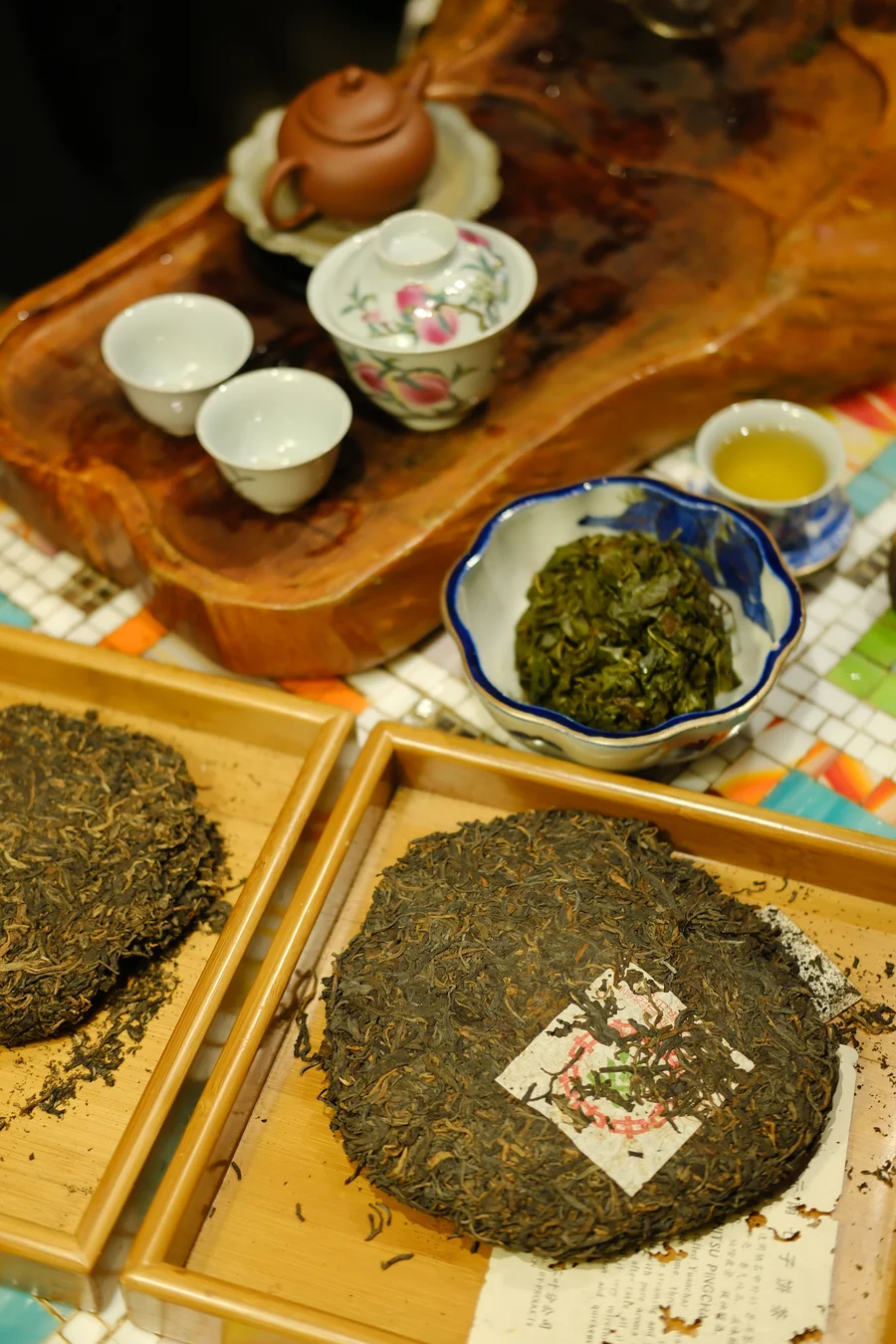 Wooden trays of large round pressed tea cakes beside a bowl of loose green leaves